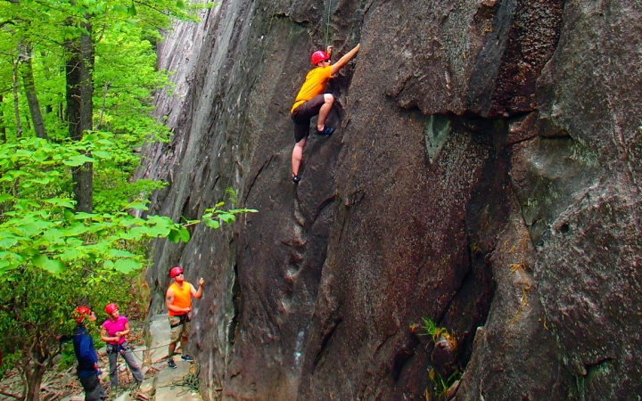 A person wearing safety gear is secured by ropes as they climb a rock wall. On the ground, one person belays while others look on. 