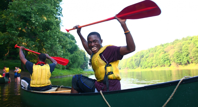 Two young people wearing life jackets sit in canoes and raise their paddles into the air in apparent celebration. One of them smiles at the camera. 