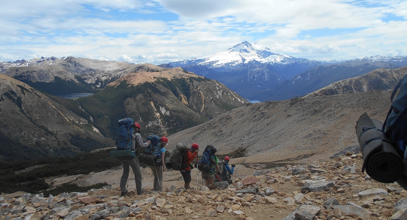 A group of people wearing backpacks hike along a rocky landscape. There is a vast mountainous landscape in the distance.
