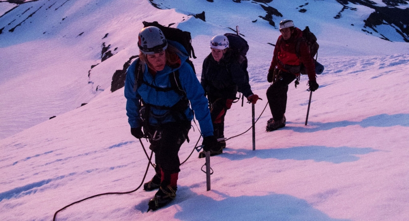 A group of outward bound students wearing mountaineering gear make their way up a snowy incline. The snow appears pink from the sunrise or sunset. 