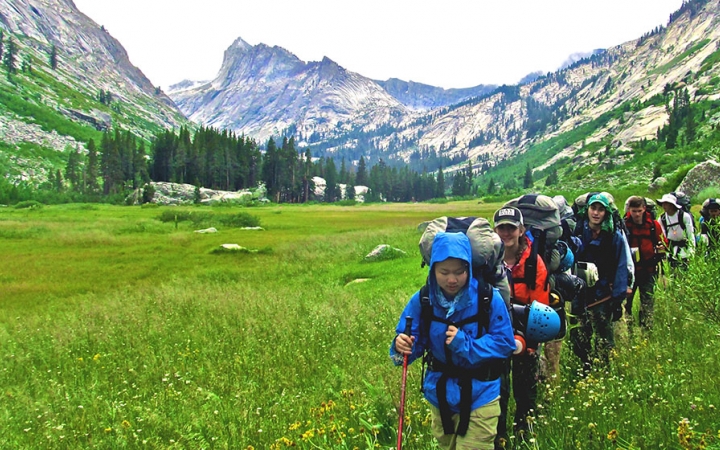 A group of young people wearing backpacks hike through a valley of bright green grass. There are mountains and evergreen trees in the background.