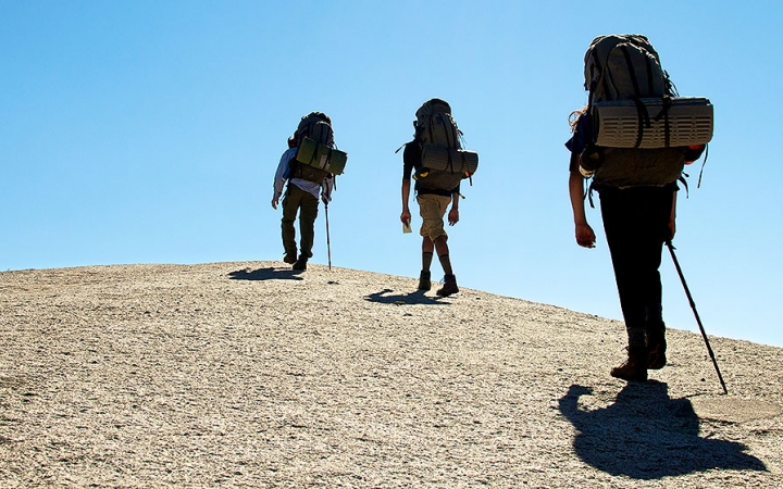 Three people wearing backpacks hike away from the camera up a rock incline. There is a blue sky above.