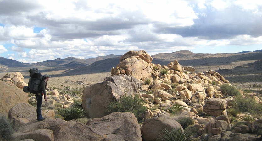 A person wearing a backpack looks out over a rocky landscape. There are mountains in the distance. 