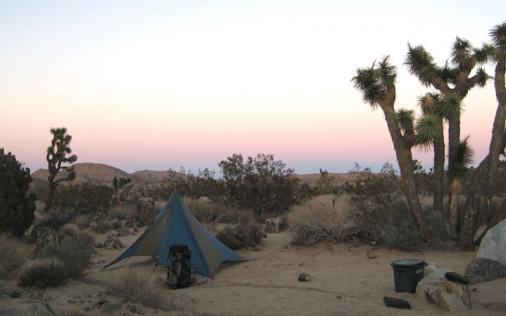The sun rises or sets behind a campsite in Joshua Tree National park. The sky appears in colors of blue, yellow, pink and purple. 