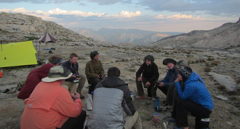 A group of outward bound students sit in a circle amidst a rocky landscape. Their tents sit nearby. There are mountains in the background. 