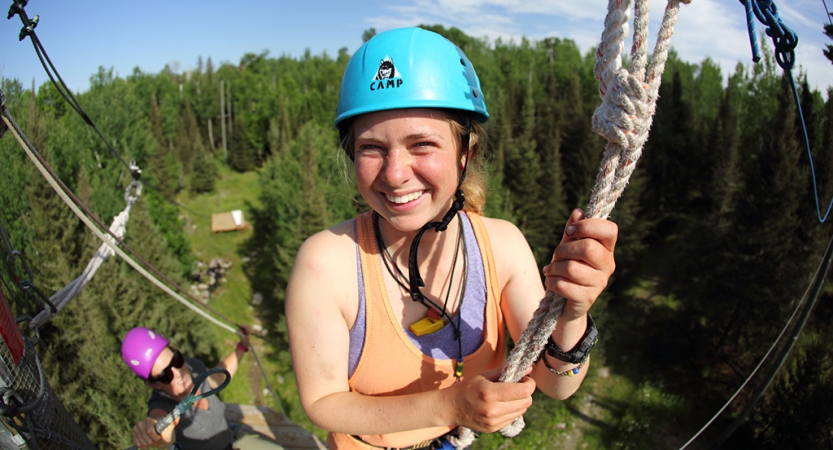 A person wearing safety gear and secured by ropes smiles as they make their way through a high ropes course.
